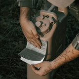 A close-up of a person's hands applying an instruction sheet to a beige plastic jerrycan outdoors on grass.