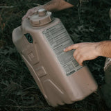 A person outdoors holds a large beige plastic water container, pointing to printed instructions on the side.