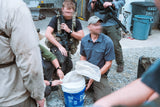 Person in tactical gear interacting with another person in a blue shirt outdoors.