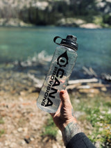 Hand holding a clear water bottle with 'Cana Provisions' branding against a natural background.