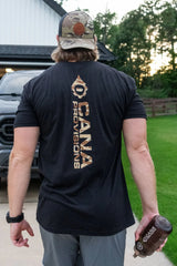 A man viewed from behind wearing a black t-shirt with a large camo vertical 'CANA PROVISIONS' logo on the back, paired withg rey shorts and a camo baseball cap, standing outdoors on a paved path with trees in the background.
