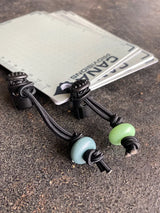 one blue lumimod glow washer and one green lumimod glow washer attached to draw strings that are attached to cana provisions water data cards, on a concrete surface