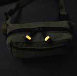 An olive green fanny pack with black zippers and bright orange glowing beads on the front, shown against a dark background.