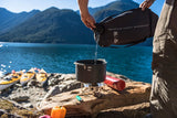 Person using a MSR 6l dromedary water reservoir bag pouring water into a cooking pot by a lake with mountains in the background