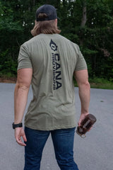 A man viewed from behind wearing an olive green t-shirt with a large black vertical 'CANA PROVISIONS' logo on the back, paired with blue jeans and a black baseball cap, standing outdoors on a paved path with trees in the background.