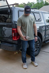 A man wearing a black cap, a green t-shirt, and blue jeans stands in front of a dark gray pickup truck, holding a water bottle