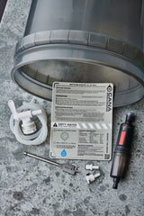 Water filtration system components including a bucket, valve, drill bit, and tools on a concrete surface.