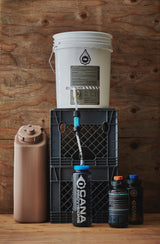 Collection of Cana Provisions products including a white bucket, brown jerry can jug, black crate, and black bottles on a wooden surface.